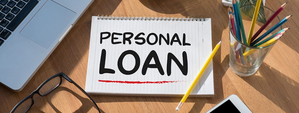 'PERSONAL LOAN' written on a notebook beside laptop and coloured pencils in a glass tumbler